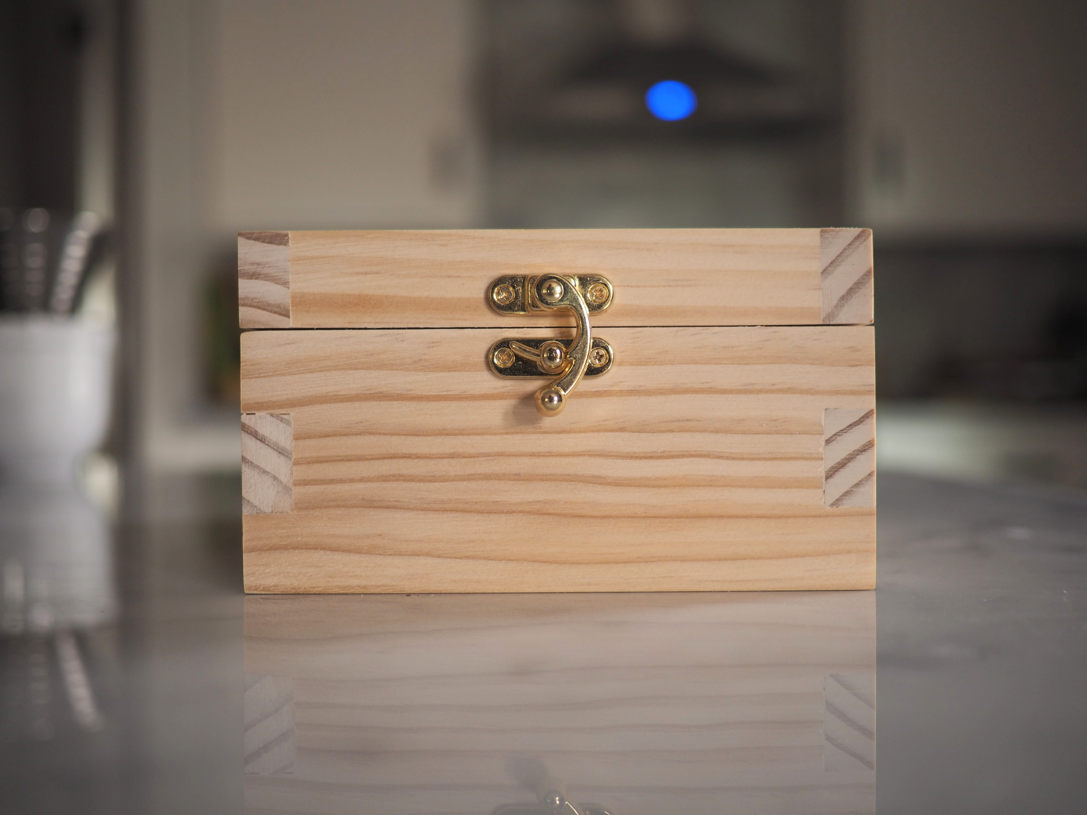 A front-on view of the box showing the contrasting grain direction of the box joints and the semicircular brass latch. The box is reflected in the marble countertop.