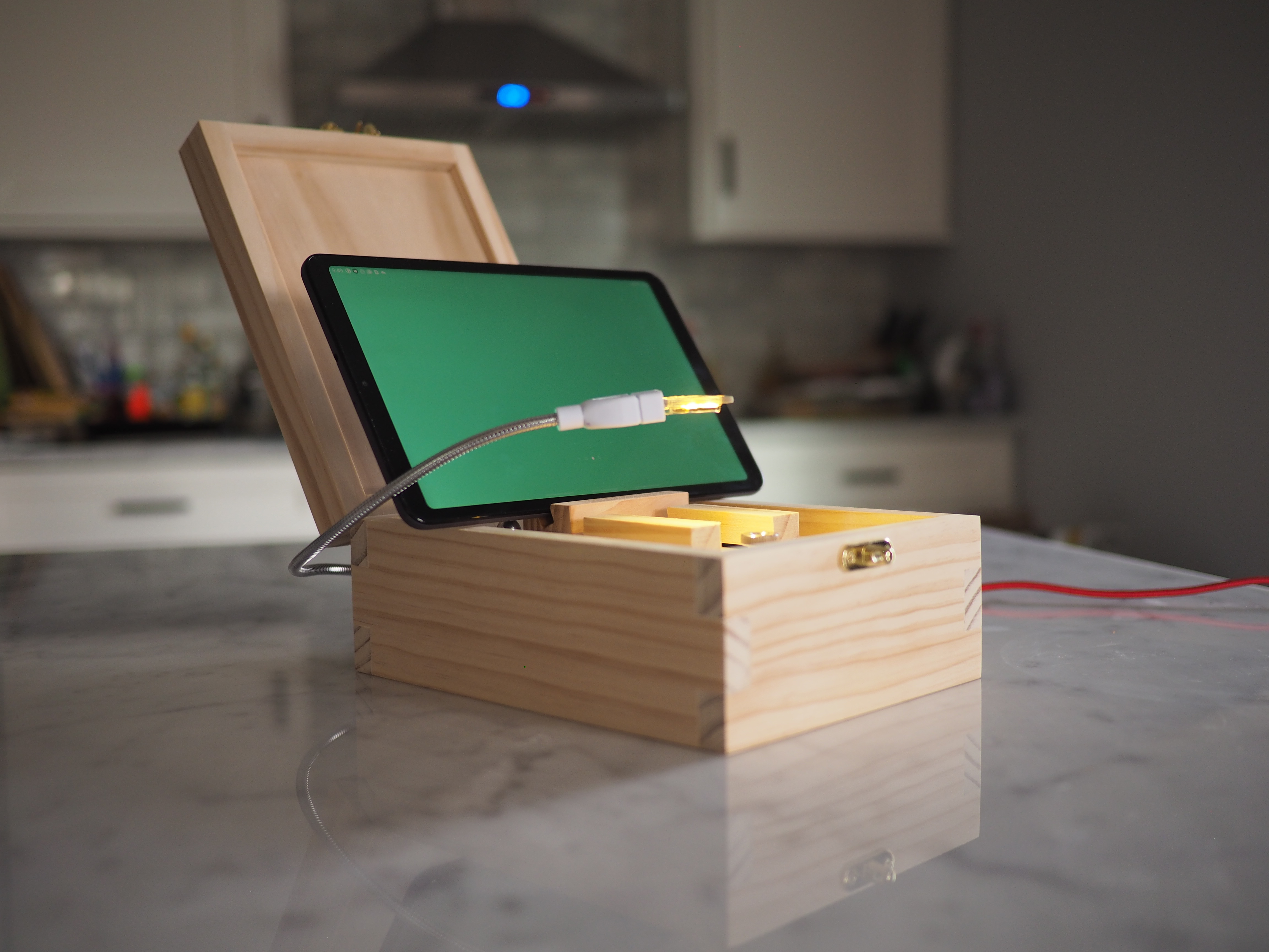 A small pine box with interlocking joints sits on a marble countertop in a dimly lit room, lid open, with a green-screened tablet resting on top and a yellow light illuminating the box interior from a silver gooseneck hovering above it, which leads to the rear of the box. A red cable runs off the table in the background.