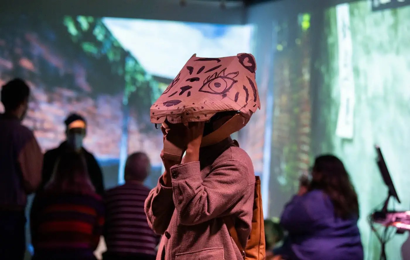 A visitor wears a cardboard tiger head mask, holding it steady by reaching into its mouth, while a group in the background sits looking at a projected 3D scene of brick walls and vegetation.