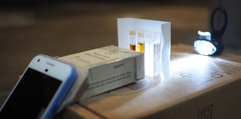 a smartphone taped to the spectrometer, pointed at three small plastic containers filled with different shades of brown liquids, backlit